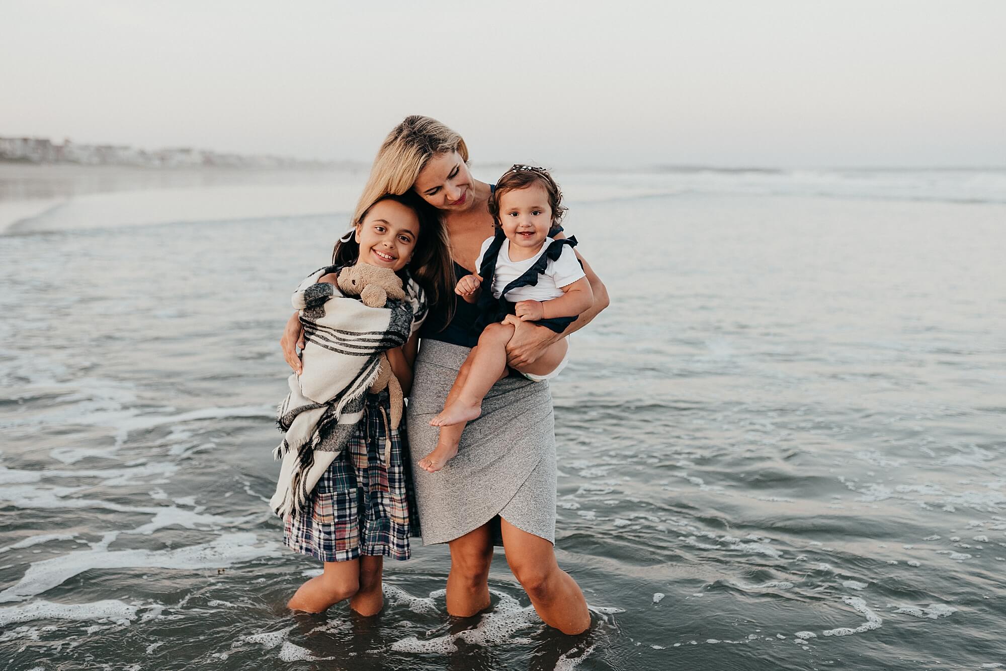 A mother holds her toddler and hugs her oldest daughter on the beach during their cape cod family photographer session