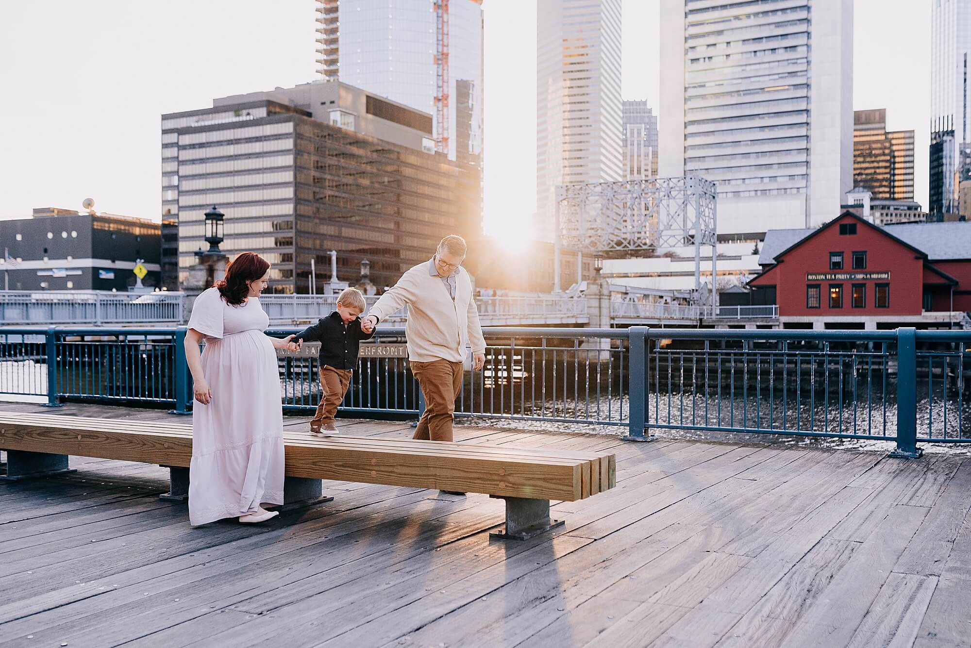 A mother and father hold their young son's hand as he walks on a bench during their Boston Maternity Photographer session