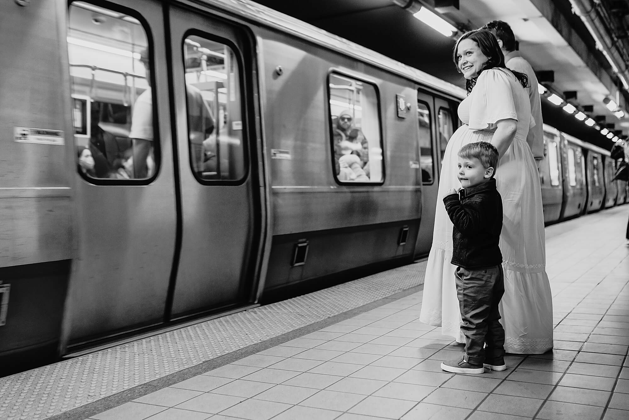 A young boy hold's his pregnant mother's hand as they wait for the train during their boston maternity photographer session