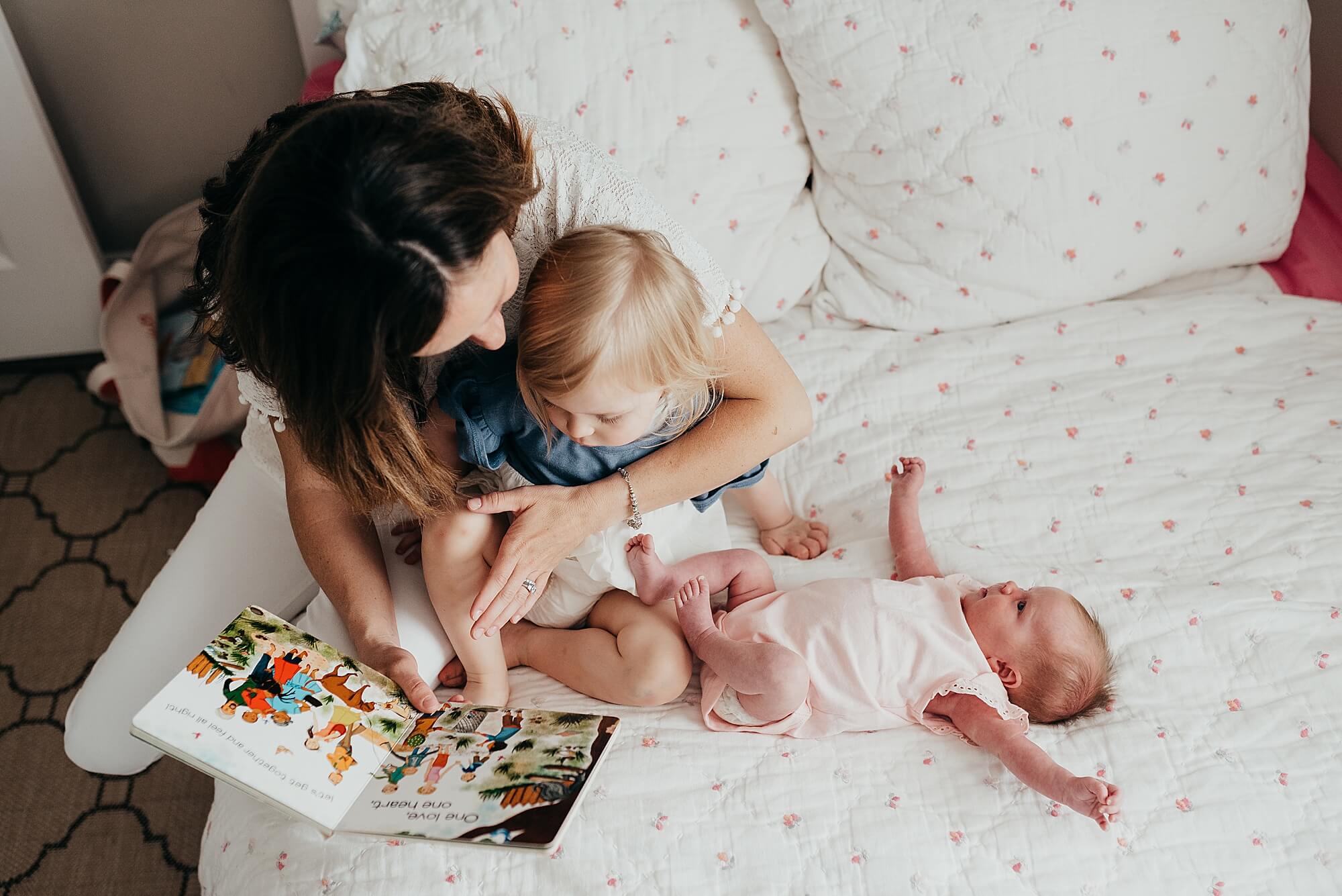 A mother reads to her toddler on the bed with her newborn baby