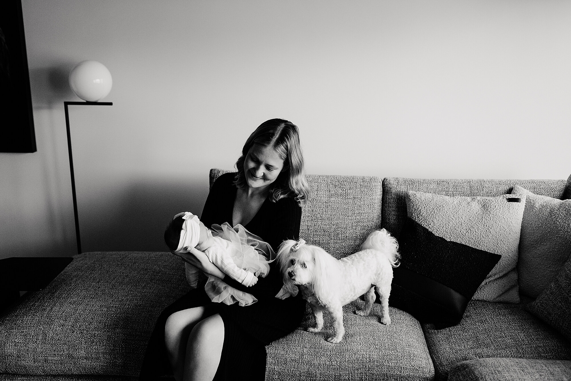 A black and white image of a mother sitting on a couch with her newborn daughter and cute little dog