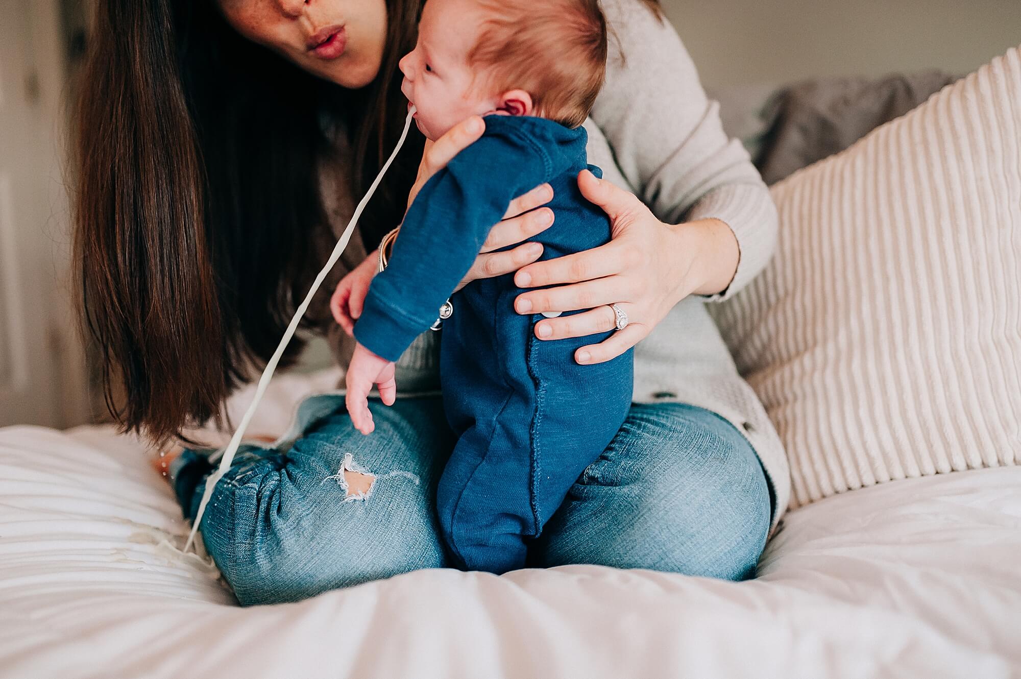 A baby spitting up on the bed, his mother can be seen behind him looking surprised