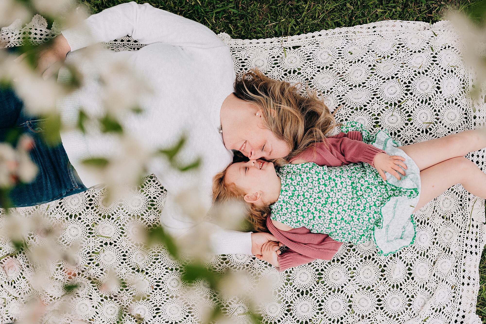 A mother lays on a blanket under a blossoming tree with her young daughter