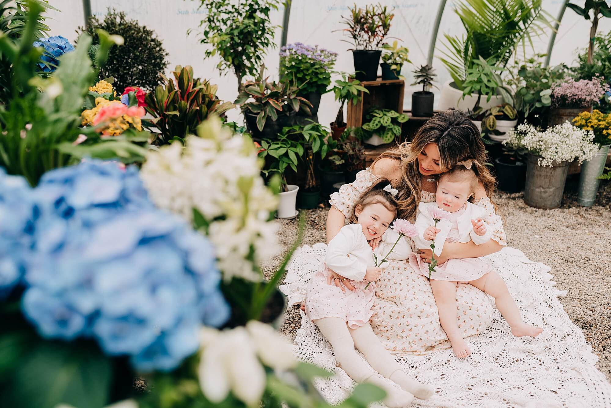 A mother snuggles her daughters in a greenhouse during a minis session with Scituate Photographer, Allison Wolf