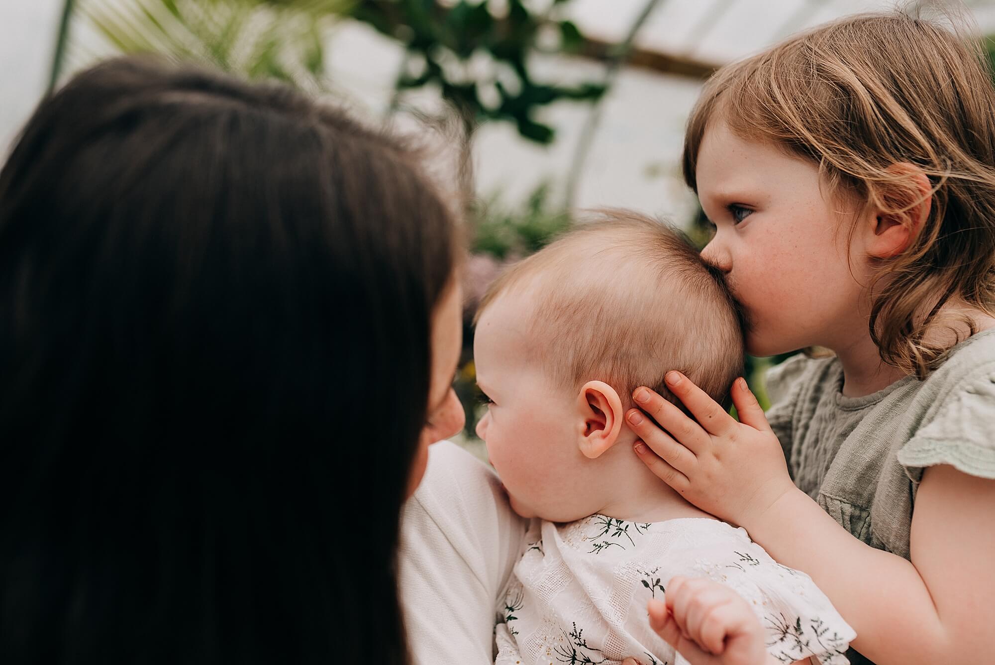 A mother holds her young daughters, the oldest gives the baby a kiss on the back of her head